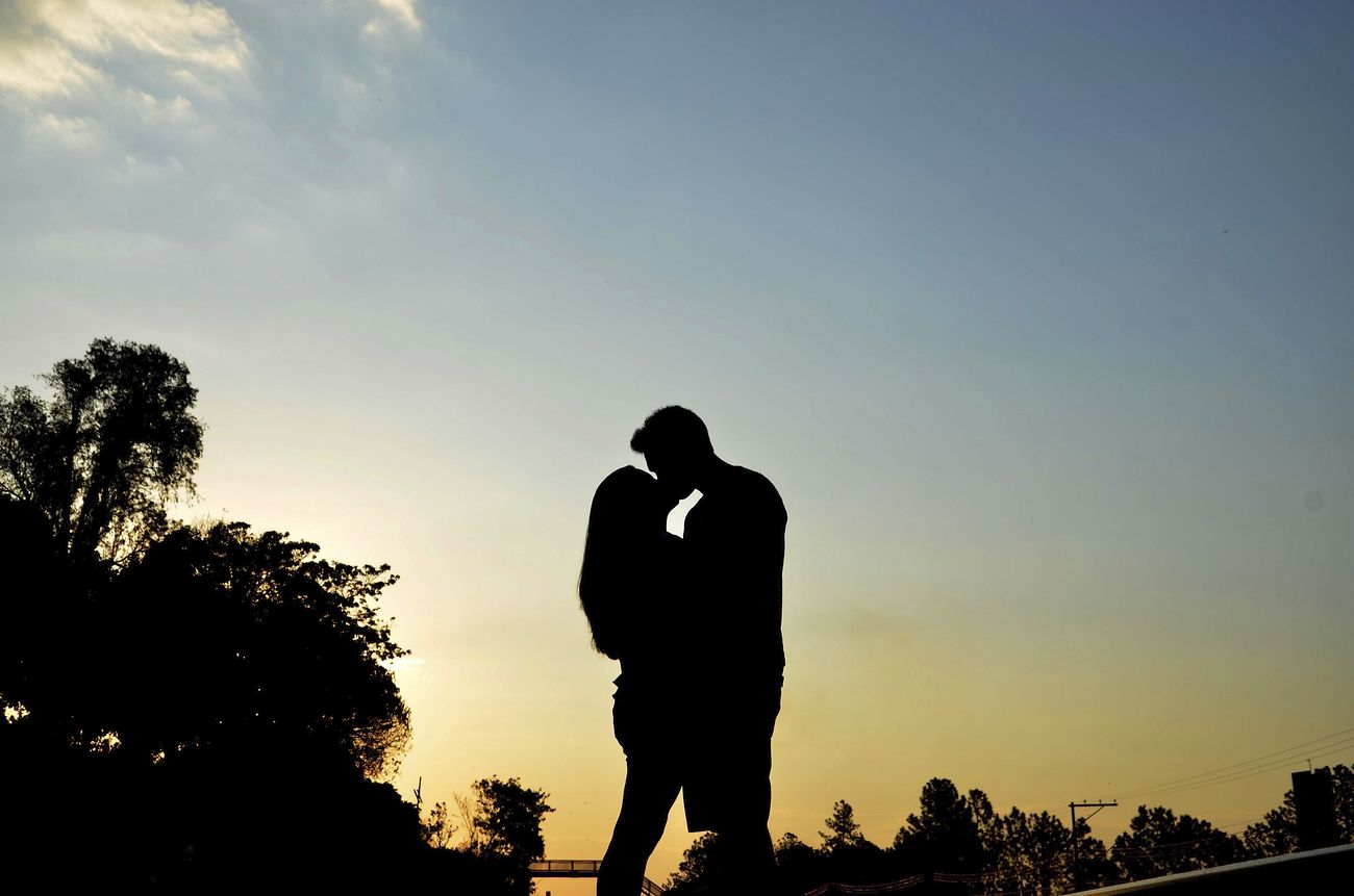 The image shows the silhouette of a man and woman kissing against a sunset with trees and a bridge in the background.