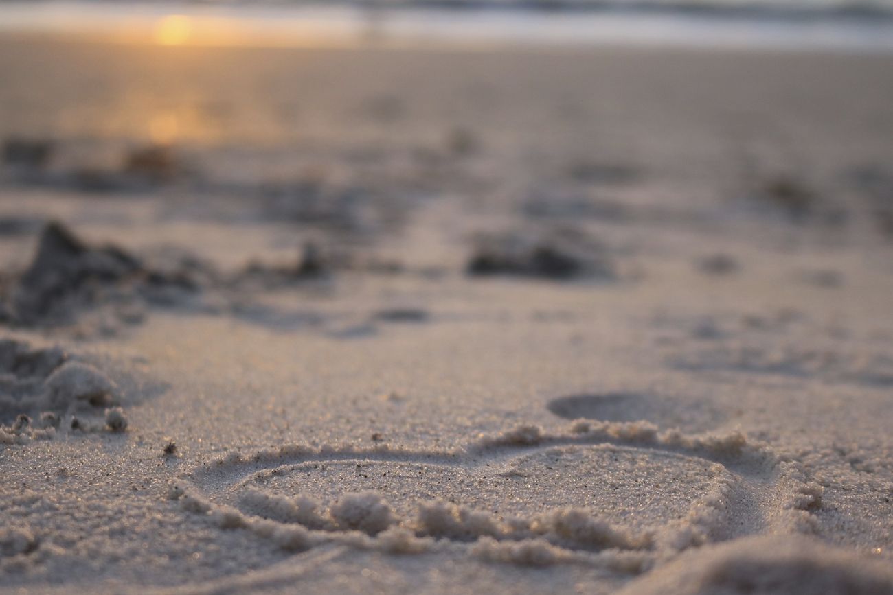 Image shows a heart drawn in the sand with the sun reflected in the sea in the background.