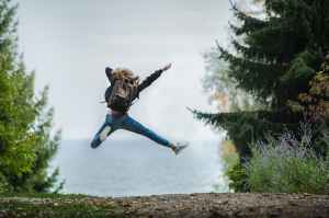 Photo shows a person wearing a backpack facing away from the camera on a woodland path, leaping in the air with their right arm and leg pointed.